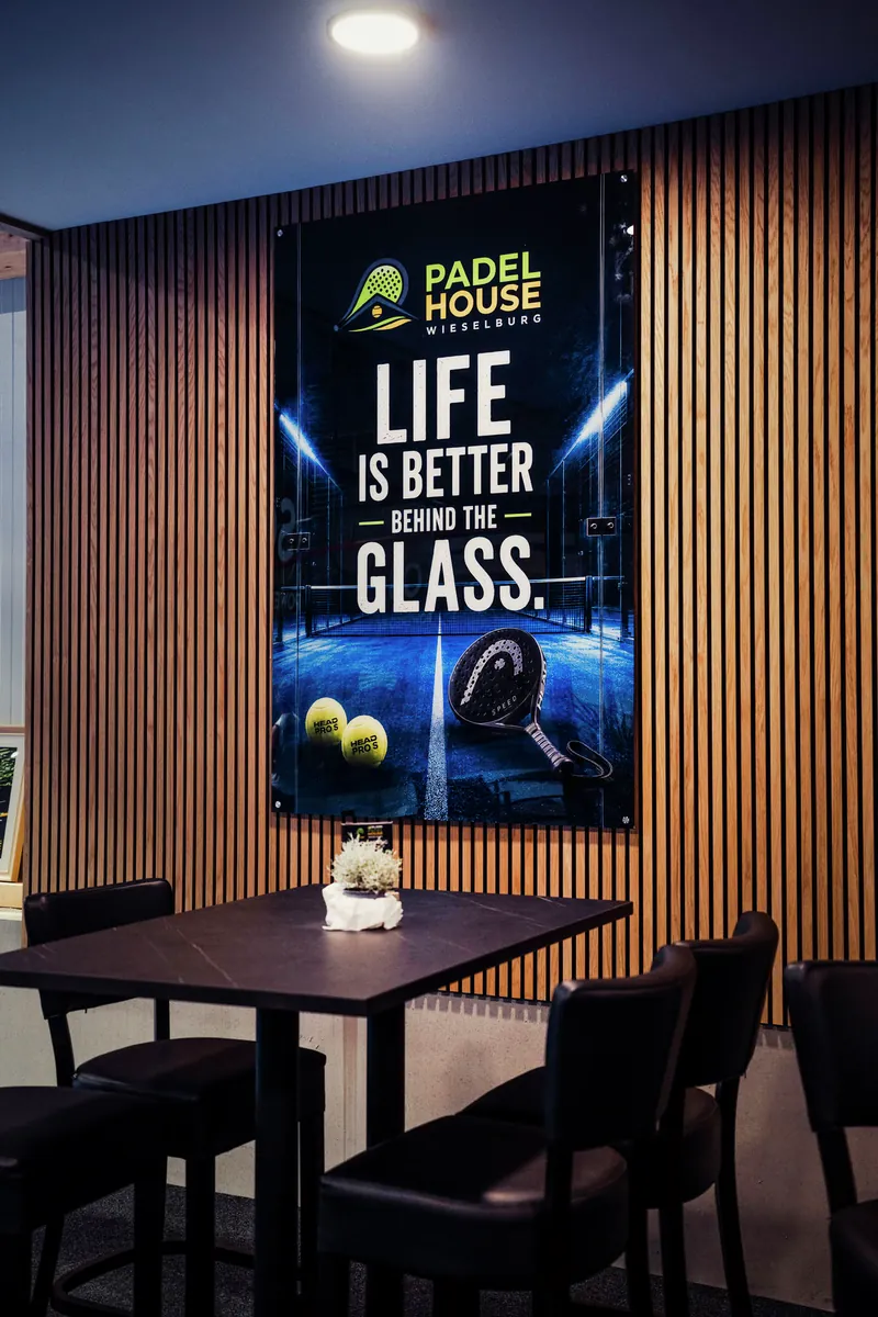 Lounge im Padelhouse: Hochtisch mit Barstühlen vor einer Holzlamellenwand mit Padelhouse-Plakat